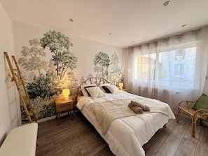 A bedroom with a large bed, a nightstand with a lamp, a chair, and a wall mural. - Vrbo Property (Vesoul)