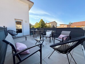 En udendørs terrasse med siddepladser og et bord, udsigt over kvarteret og en klar himmel.