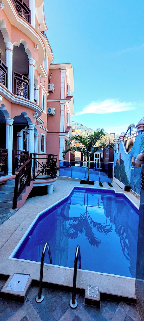 A swimming pool with a clear blue water surface, surrounded by a building with balconies and a decorative wall.