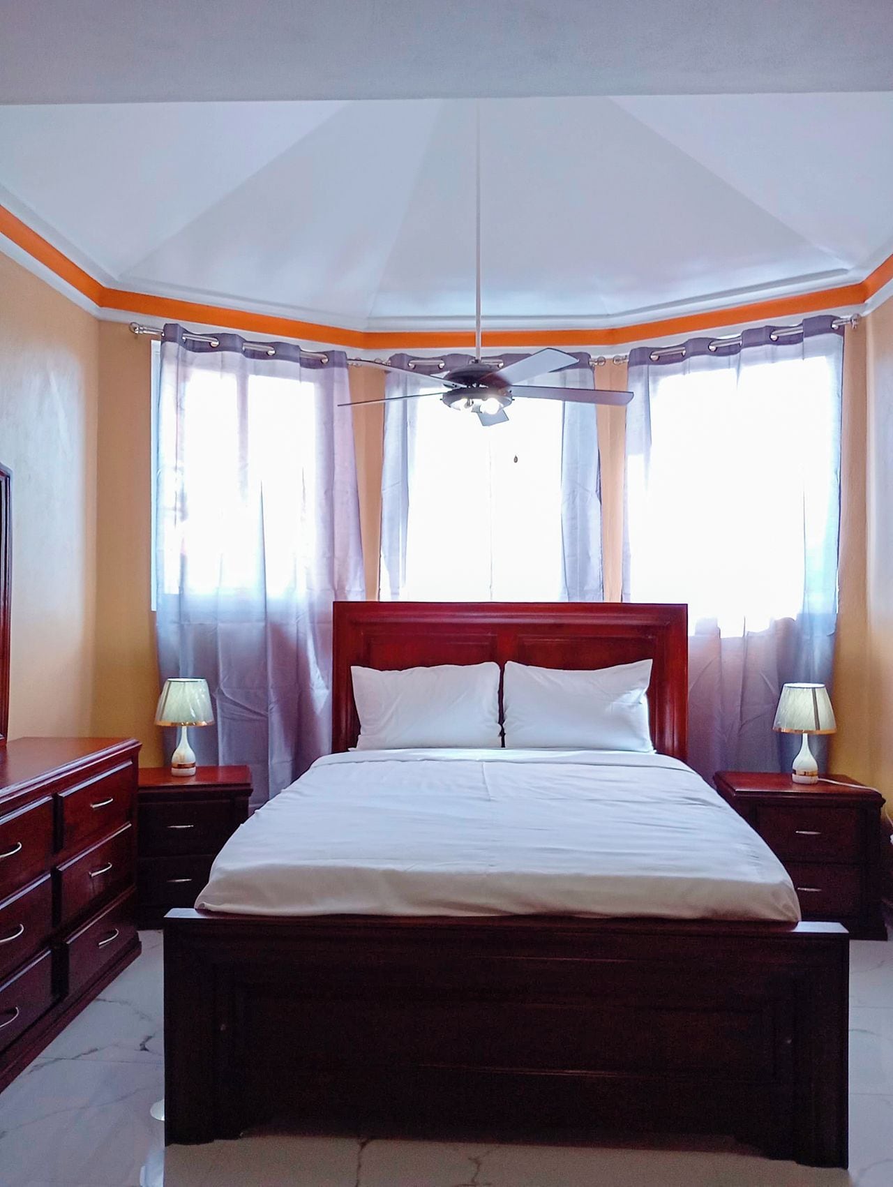 Quarto de hotel com uma cama grande, duas mesas de cabeceira com abajures, uma cabeceira de madeira e um ventilador de teto.