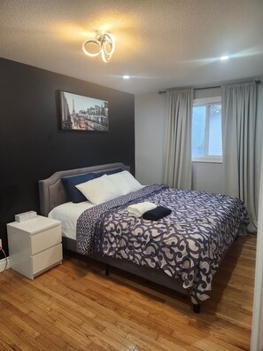A bedroom with a bed, a nightstand, a wall-mounted light fixture, a window with curtains, and a cityscape painting. - Charming & Cozy Guest House in Windsor (Windsor)
