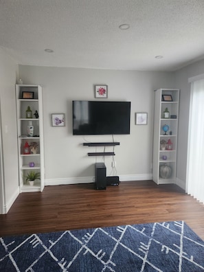 A living room with a flat-screen TV mounted on the wall, wooden flooring, and a blue patterned rug. - Charming & Cozy Guest House in Windsor (Windsor)