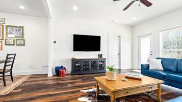 Ein geräumiges Wohnzimmer mit einem an der Wand befestigten Flachbildfernseher, einem Deckenventilator, einem blauen Sofa, einem Holztisch und einem Kuhfellteppich.