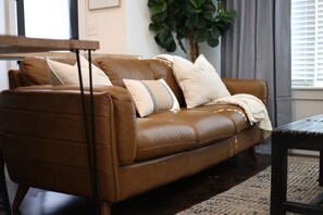 A brown leather sofa with cushions, a wooden coffee table, a large green plant, and a window with blinds. - The Fayetteville Trail House-1.5 miles from the U of A & next to Centennial Park (Fayetteville)