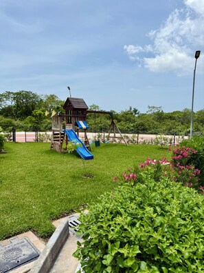 A playground with a slide and swing set, surrounded by green grass and flowering plants. - Casa Lamay | Clubhouse with a pool, internet, and a beach just a 15-minute drive away (Chuburná)