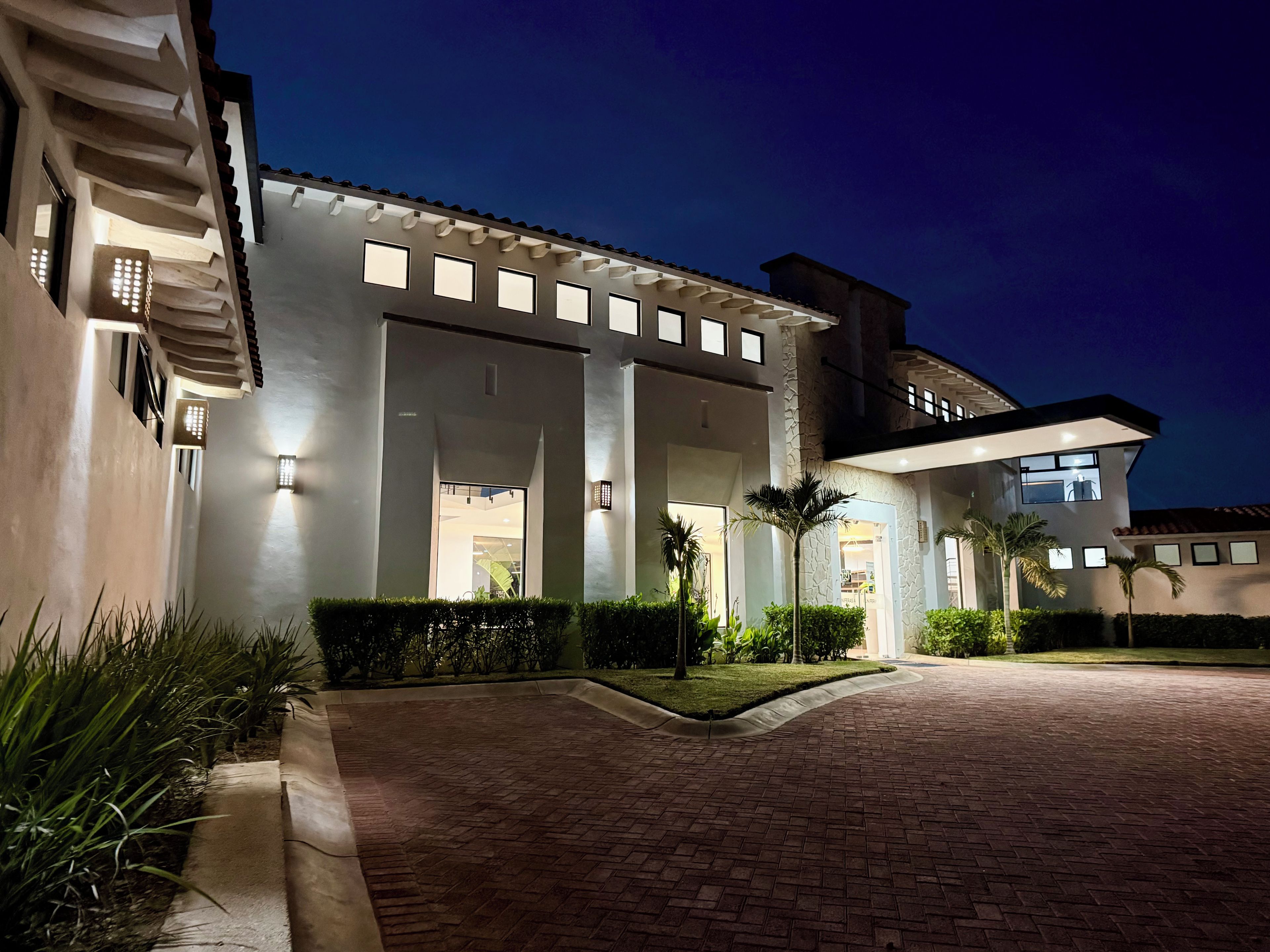 A modern building with a brick pathway leading to a gated entrance, surrounded by well-maintained greenery.