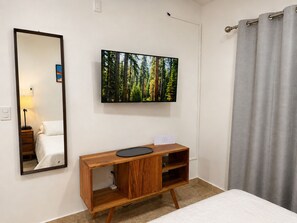 A hotel room with a bed, a wooden nightstand, a mirror, a flat-screen TV mounted on the wall, and a grey curtain. - Casa Lamay | Clubhouse with a pool, internet, and a beach just a 15-minute drive away (Chuburná)