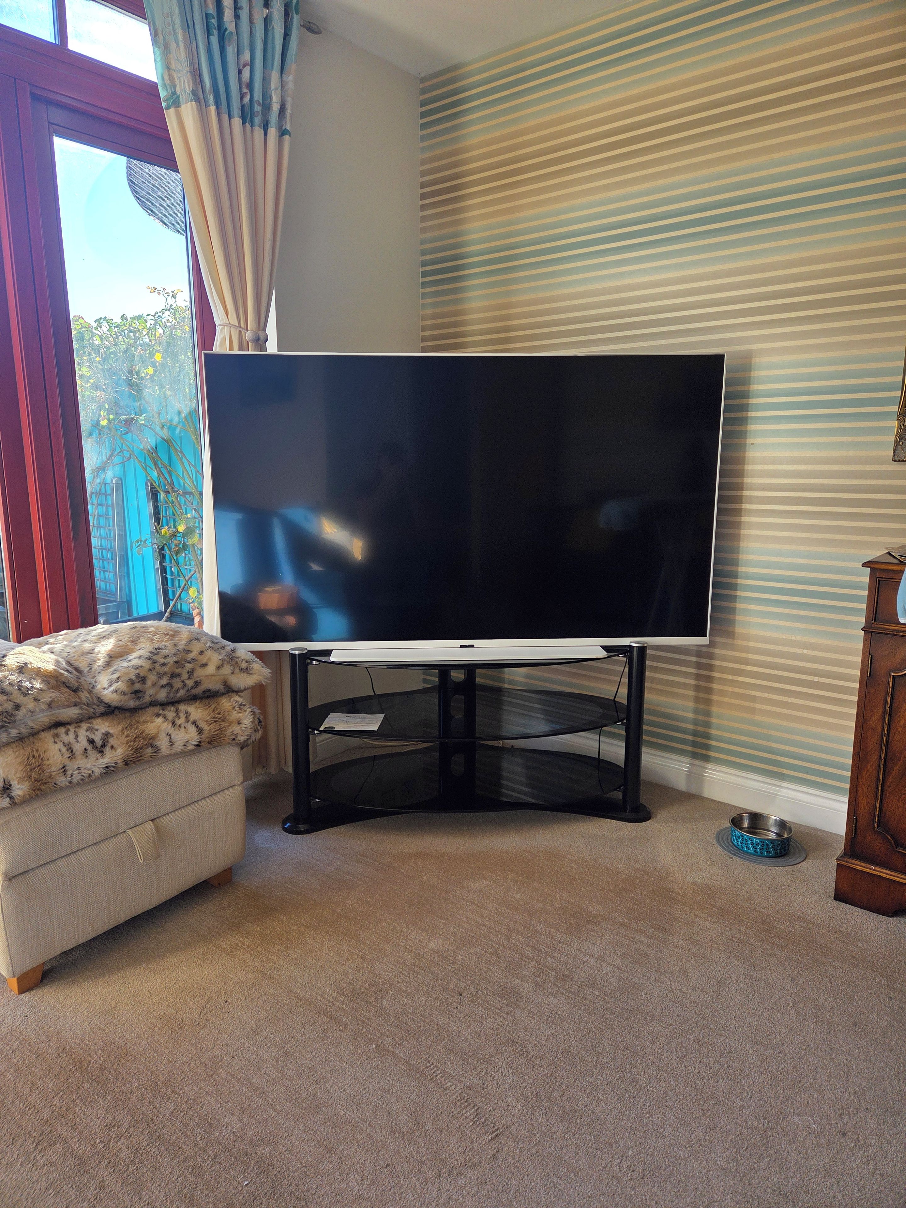 Una sala de estar con un televisor de pantalla plana grande sobre un mueble bajo, un sofá pequeño con una manta de piel, un armario de madera y una ventana con cortinas.