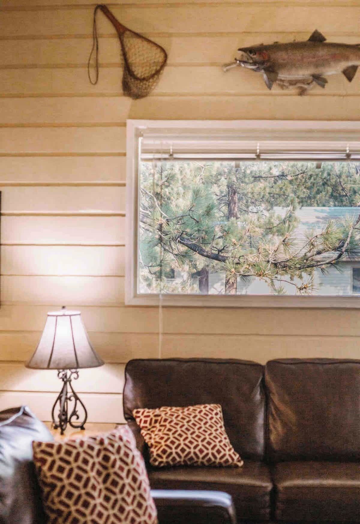 A living room with a brown leather sofa, patterned cushions, a lamp, a fish hanging on the wall, and a window with a view of trees.
