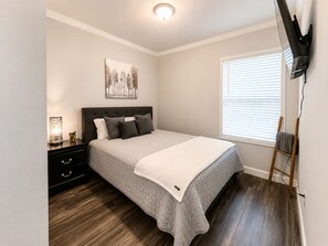 A bedroom with a bed, a nightstand, a lamp, a painting, a window with blinds, and a TV mounted on the wall. - Sweet Home | 2BR Cottage w/ Hot Tub & Fire Pit (Broken Bow)