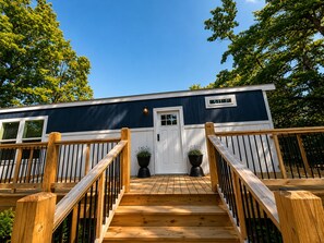 A wooden deck leading to a building with a white door and windows, surrounded by trees. - Sweet Home | 2BR Cottage w/ Hot Tub & Fire Pit (Broken Bow)