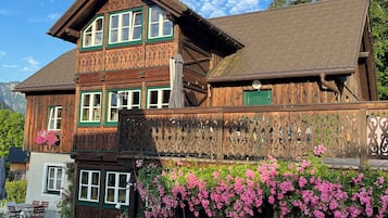 A two-story wooden house with a balcony, surrounded by lush greenery and flowering plants.