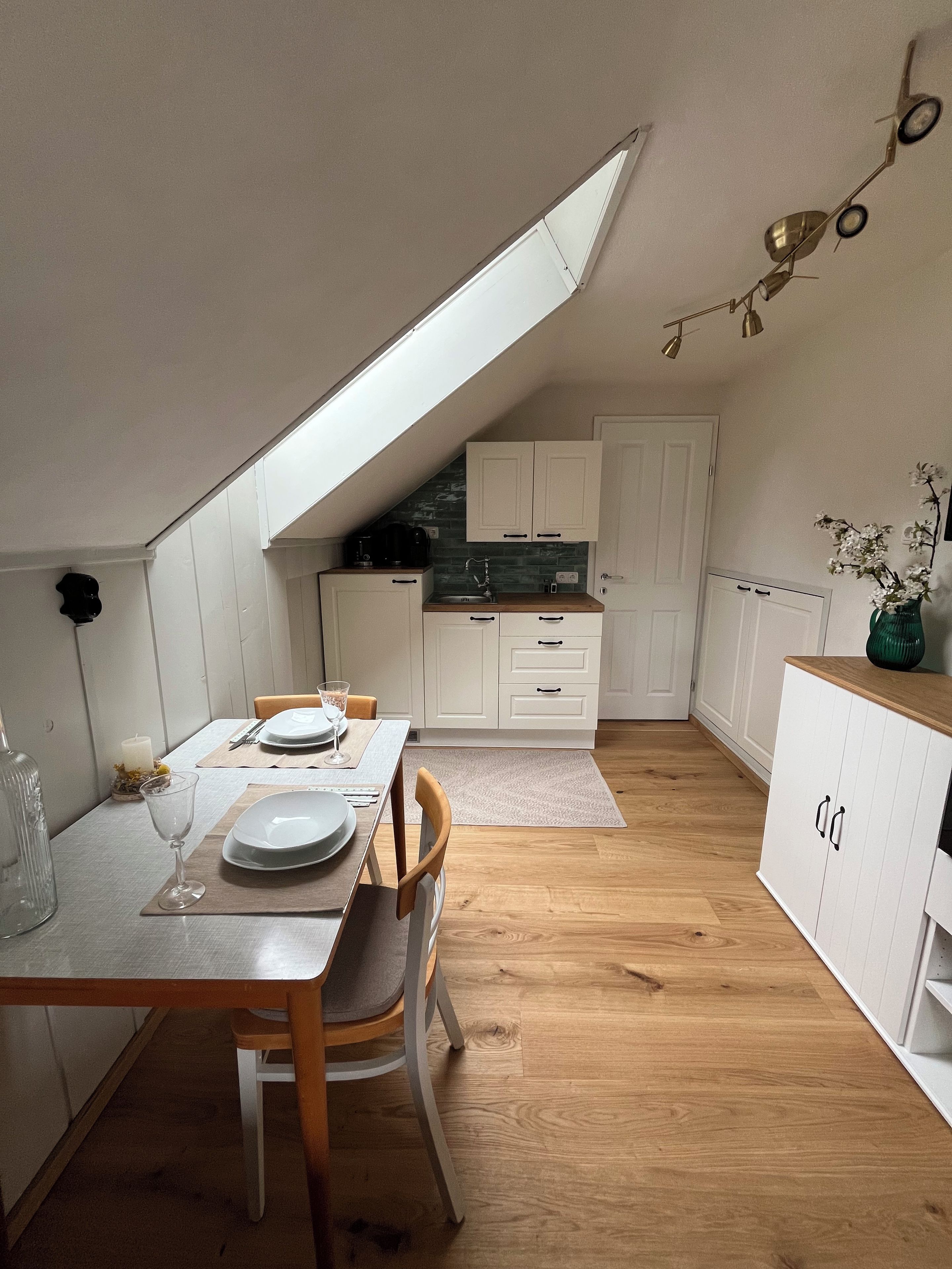 A compact kitchen with white cabinetry, a dining table set for two, and a sloped ceiling with a skylight.