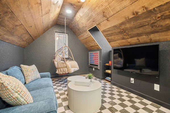 Ett mysigt rum i loftstil med ett sluttande tak, en blå soffa, en hängstol, ett runt soffbord, en tv och ett rutmönstrat golv.