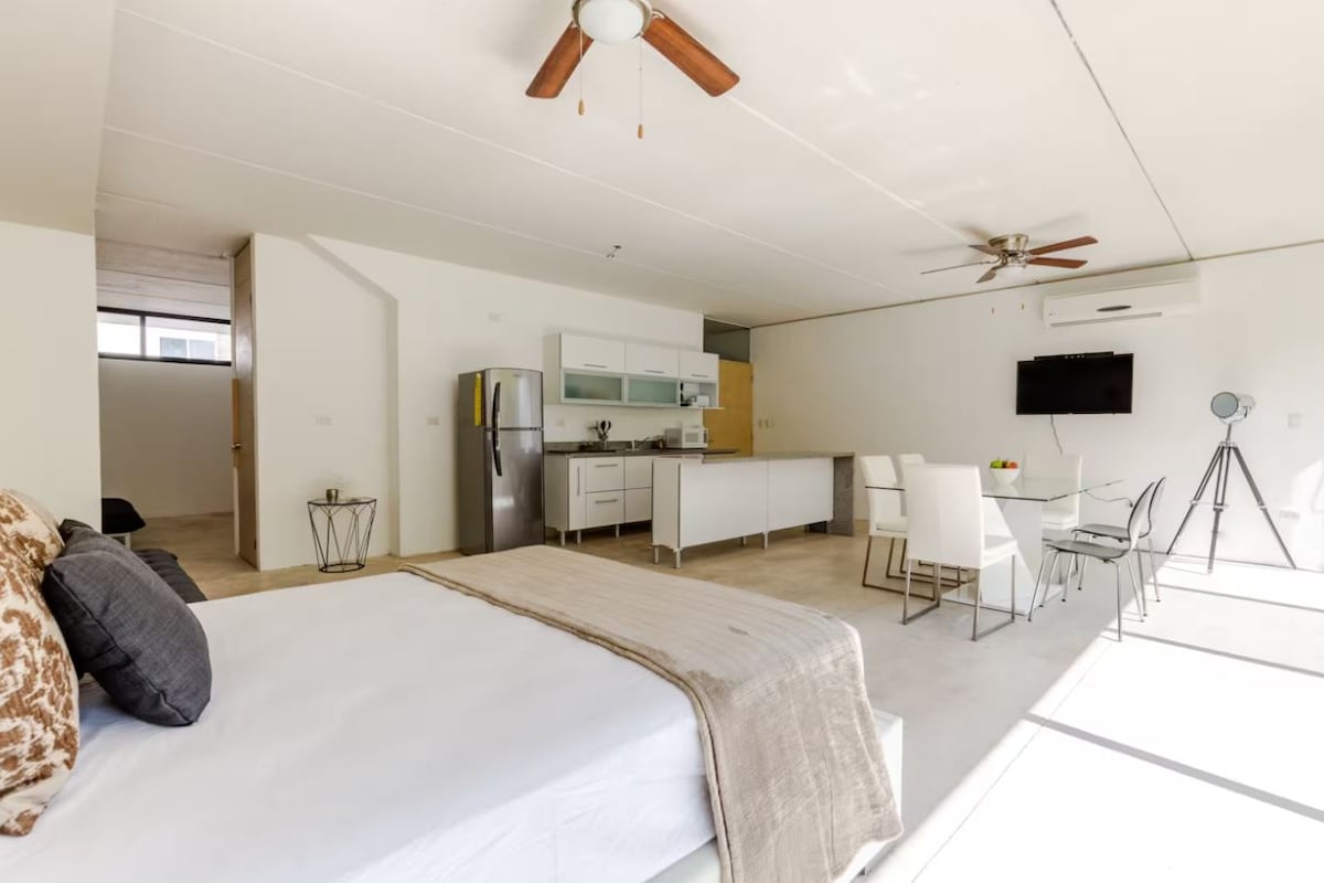 A modern bedroom with a large bed, a desk, and a kitchenette.