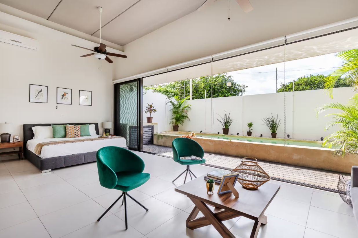 A modern bedroom with a bed, a green chair, a table, and a view of a garden.
