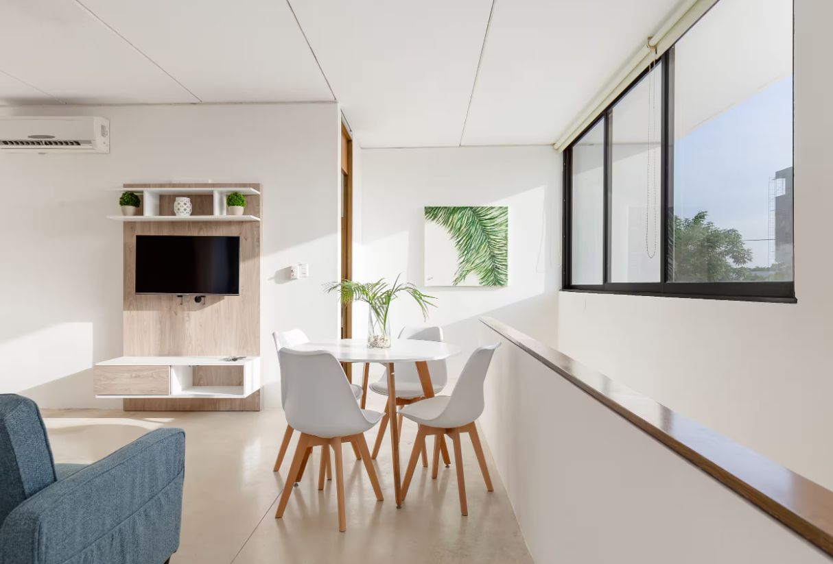 Una sala de estar moderna con un televisor de pantalla plana, un comedor con una mesa y sillas blancas, y un ventanal con vistas.