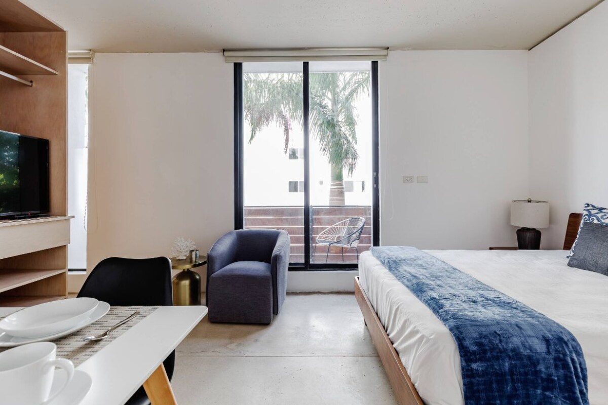 A hotel room with a bed, a desk, a chair, a television, and a balcony with a palm tree.