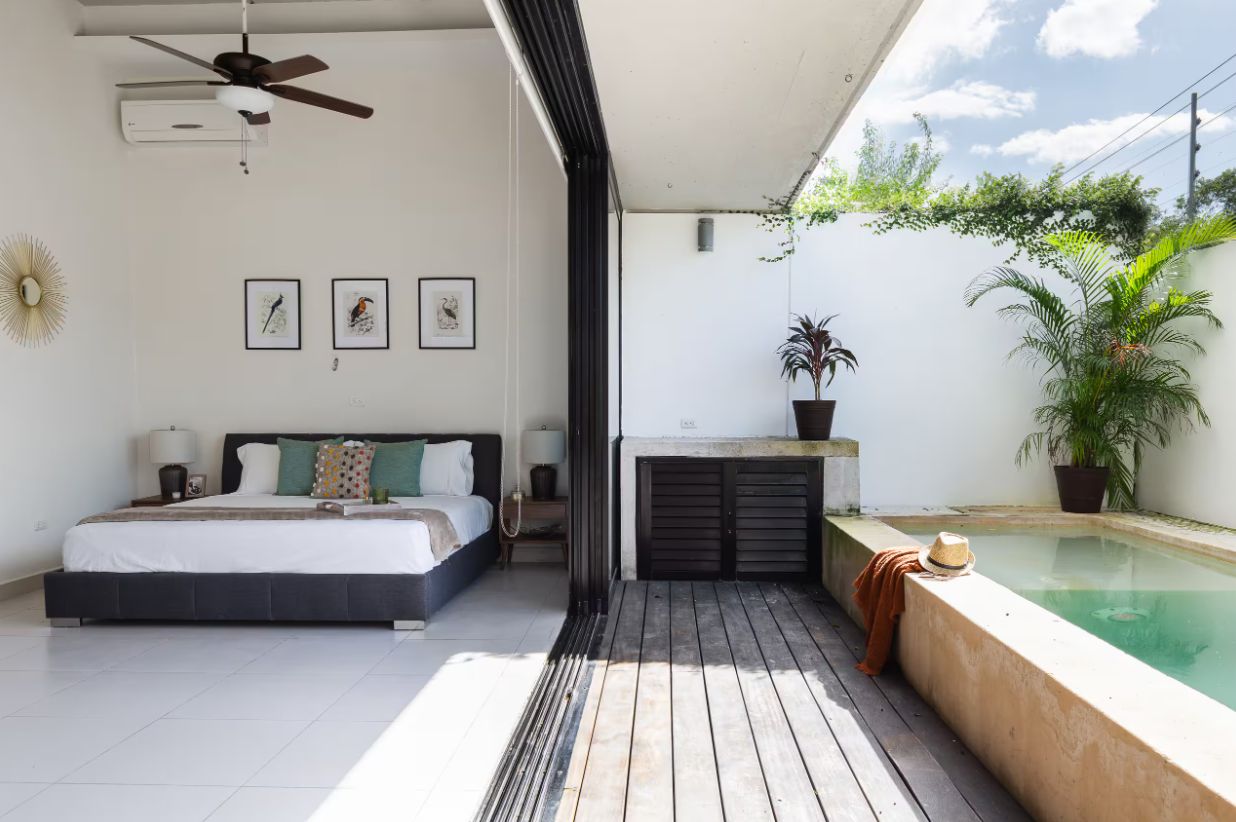 A bedroom with a bed, a ceiling fan, and a nightstand. There is a small balcony with a pool and potted plants.