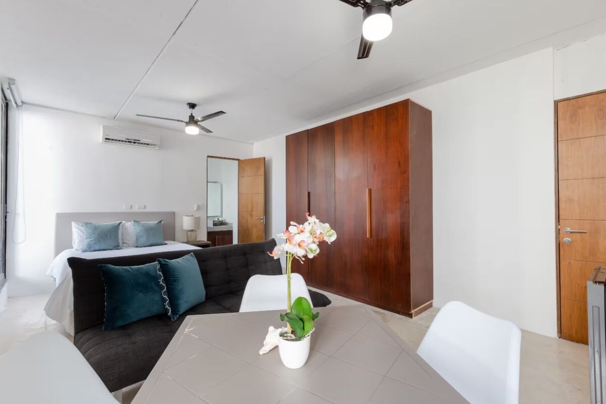 A modern hotel room with a bed, a dining table with a vase of flowers, a ceiling fan, and a wardrobe.