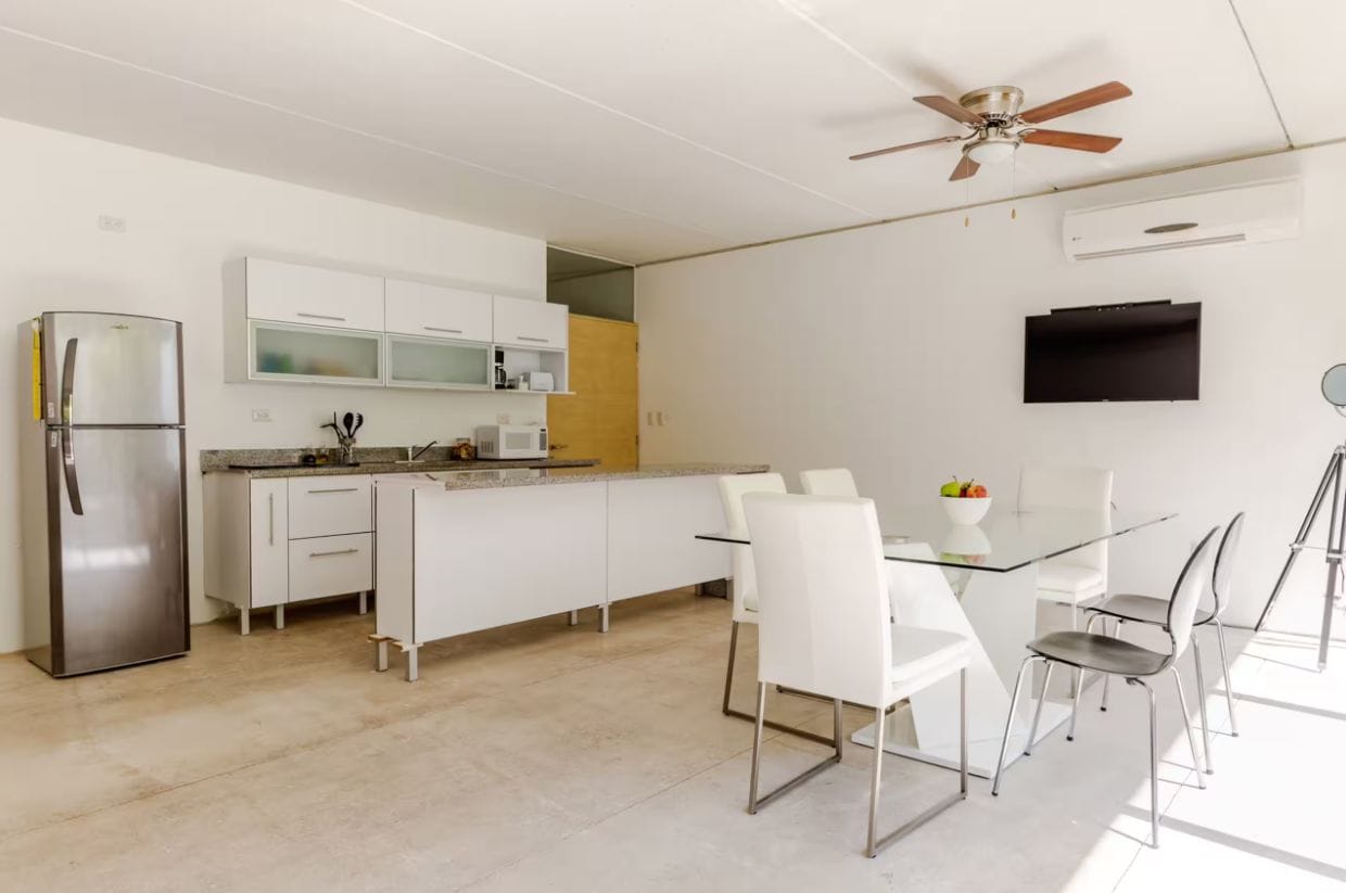 A modern kitchen with a refrigerator, microwave, and white cabinets. There is a dining table with chairs and a ceiling fan.