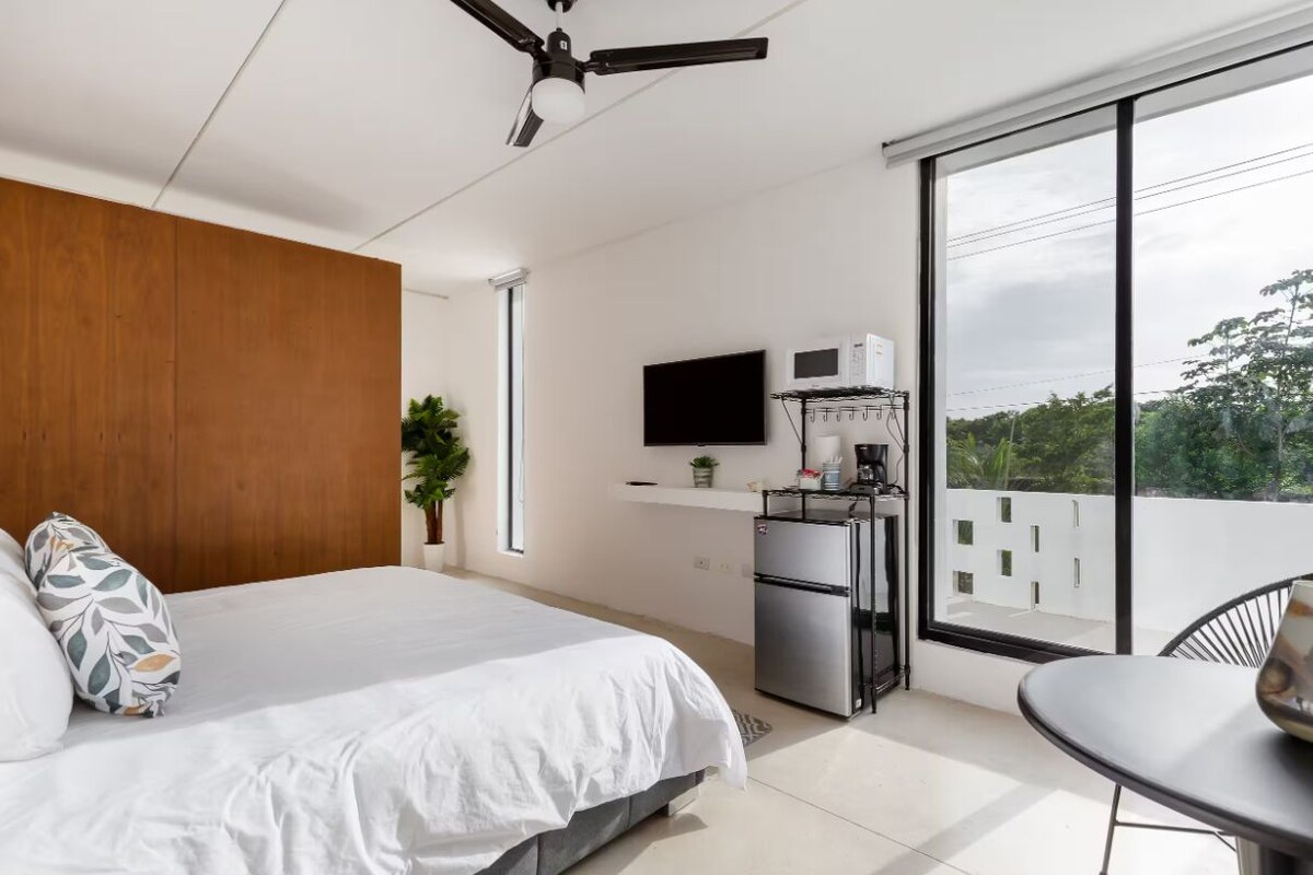 A modern bedroom with a bed, a small kitchenette, a flat-screen TV, and a balcony with a view.