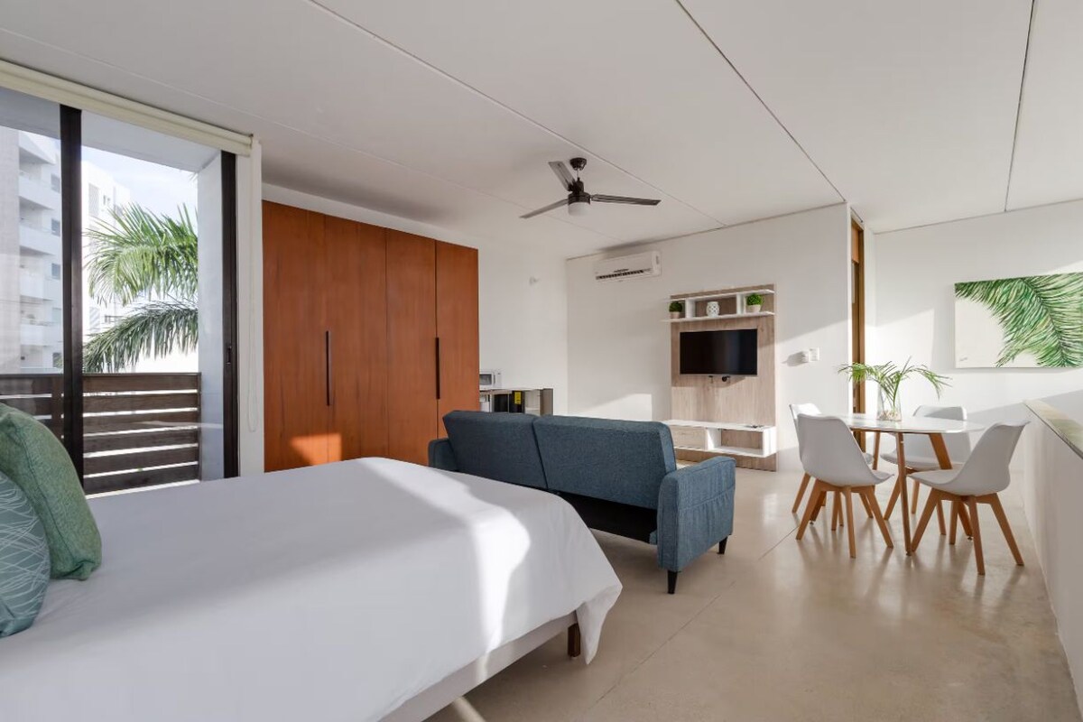 A modern room with a bed, a blue sofa, a dining area with a table and chairs, a ceiling fan, and a large window with a view of palm trees.