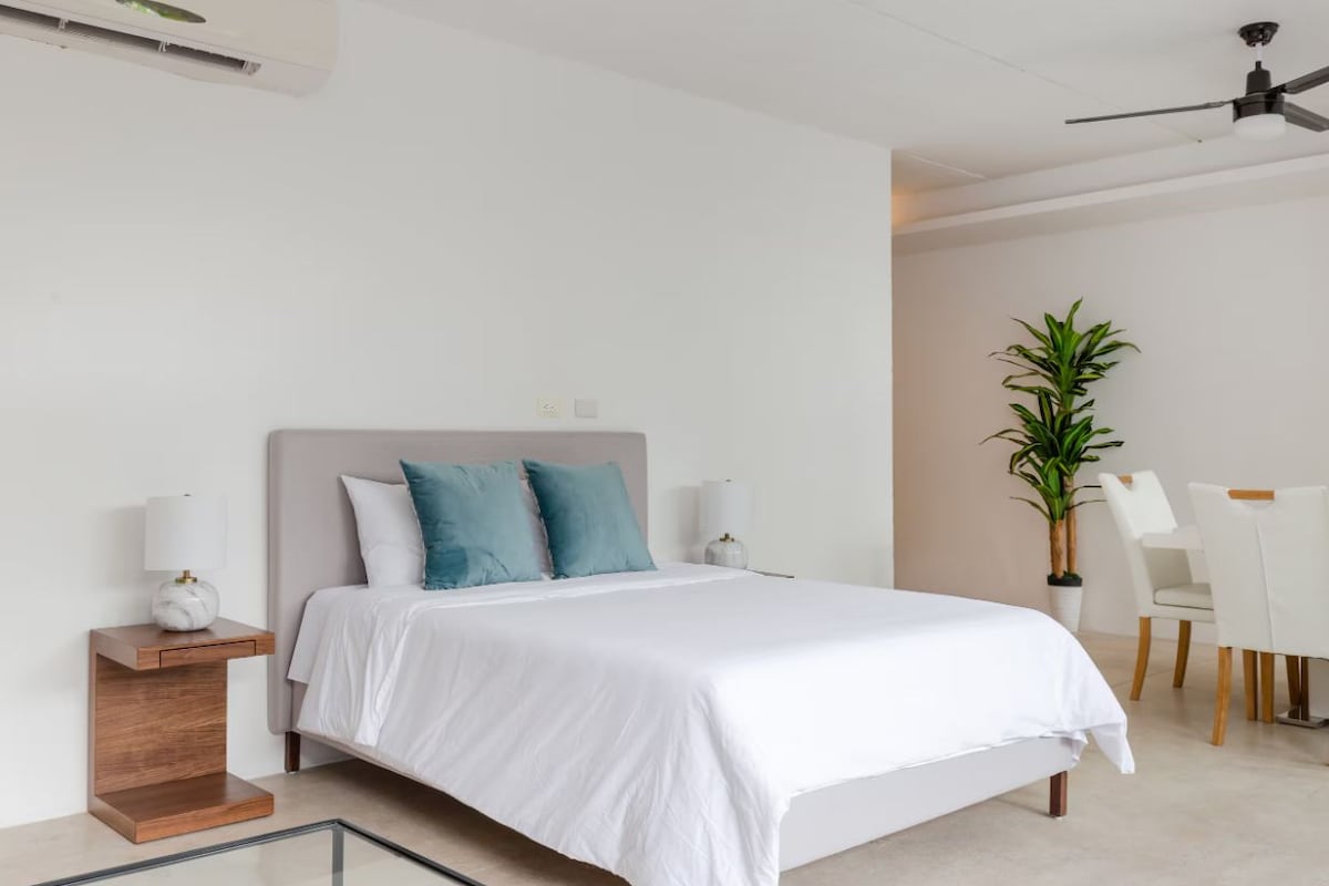 A modern bedroom with a bed, bedside tables, a plant, and a dining area with chairs.
