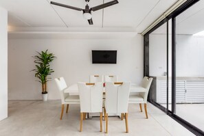 Uma área de jantar moderna com uma mesa e cadeiras brancas, um ventilador de teto, um vaso de planta e uma TV de tela plana na parede.