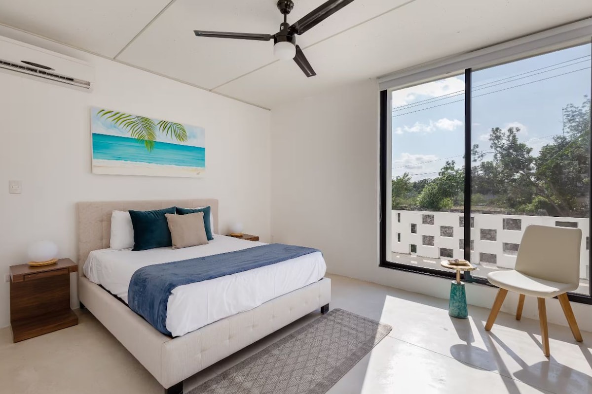 A bedroom with a bed, a ceiling fan, a window with a view, a small table, and a chair.