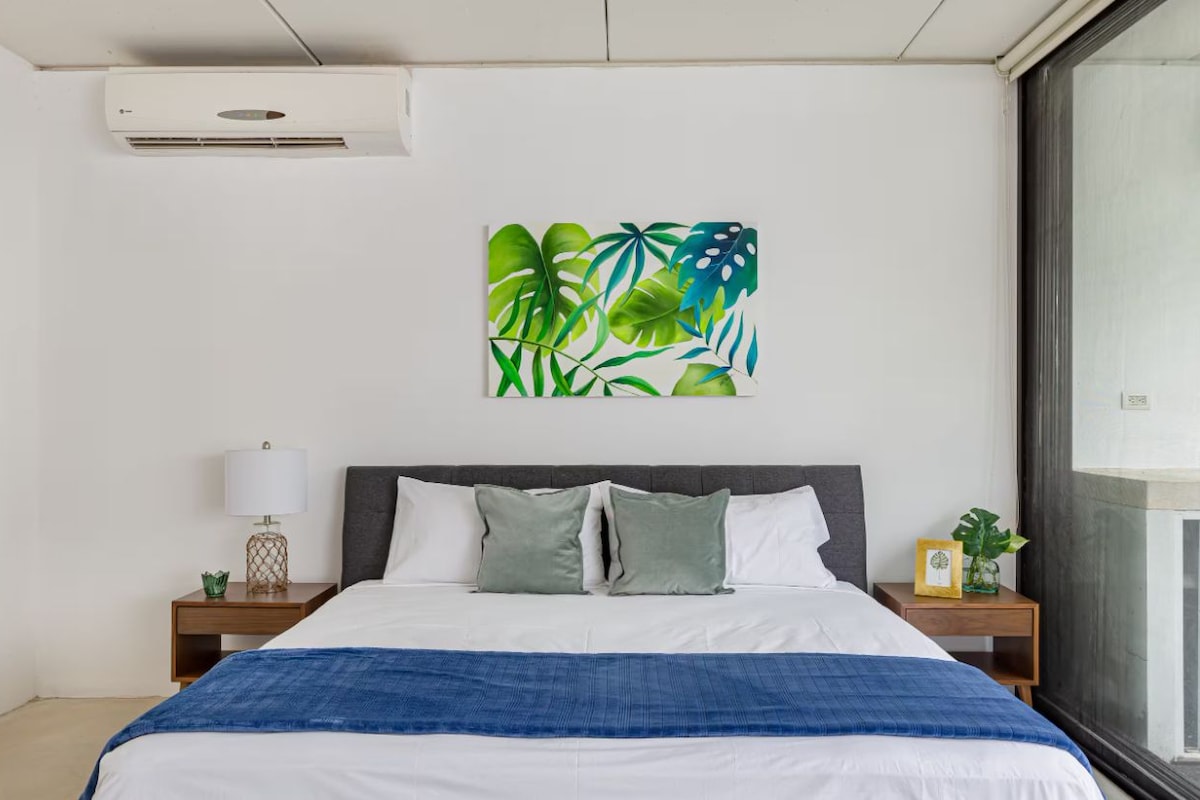 A bedroom with a bed, a nightstand, a wall-mounted air conditioner, and a window.
