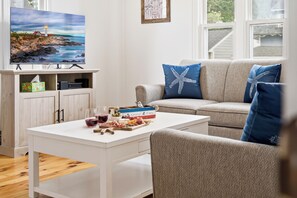 A living room with a television, a sofa with starfish pillows, a coffee table with snacks and drinks, and a wooden cabinet with a painting. - Perfect Family Beach Getaway (Old Orchard Beach)