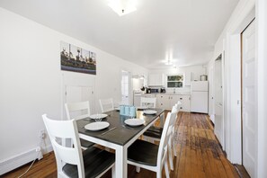 A dining area with a table set for four, white chairs, a kitchen in the background, and a door leading to another room. - Perfect Family Beach Getaway (Old Orchard Beach)