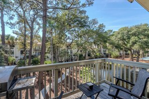 A wooden deck with outdoor furniture, surrounded by trees and a view of a residential area with multiple buildings. - Seascape Hideaway - Steps to Beach - Suite (Hilton Head Island)