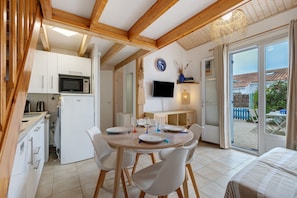 A modern kitchen with white cabinets, a microwave, and a refrigerator. There is a dining area with a table set for two, a television mounted on the wall, and a window with a view of the outside.