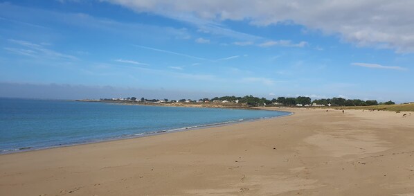 A wide sandy beach with a calm sea, a clear sky, and a distant shoreline with buildings.