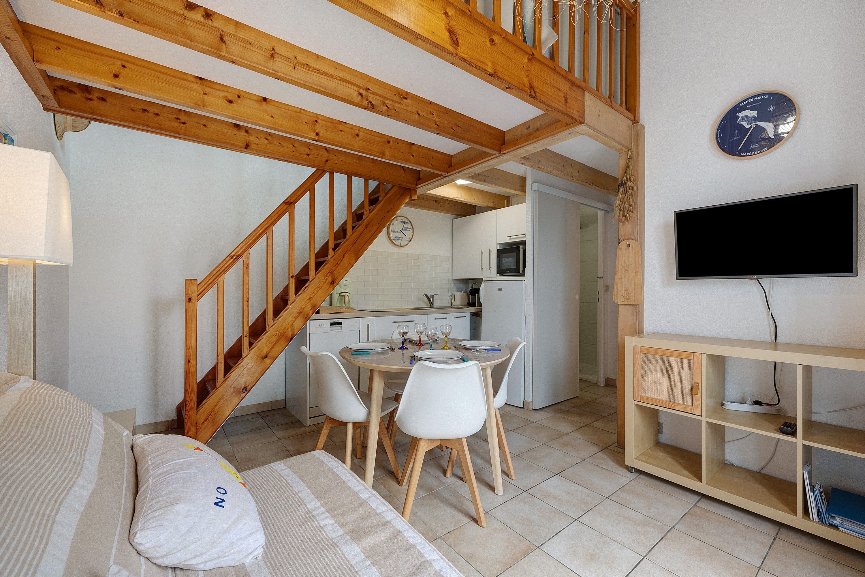 Un intérieur chaleureux avec un escalier en bois, un coin repas avec des chaises blanches, une cuisine avec des appareils électroménagers blancs et un téléviseur à écran plat fixé au mur.