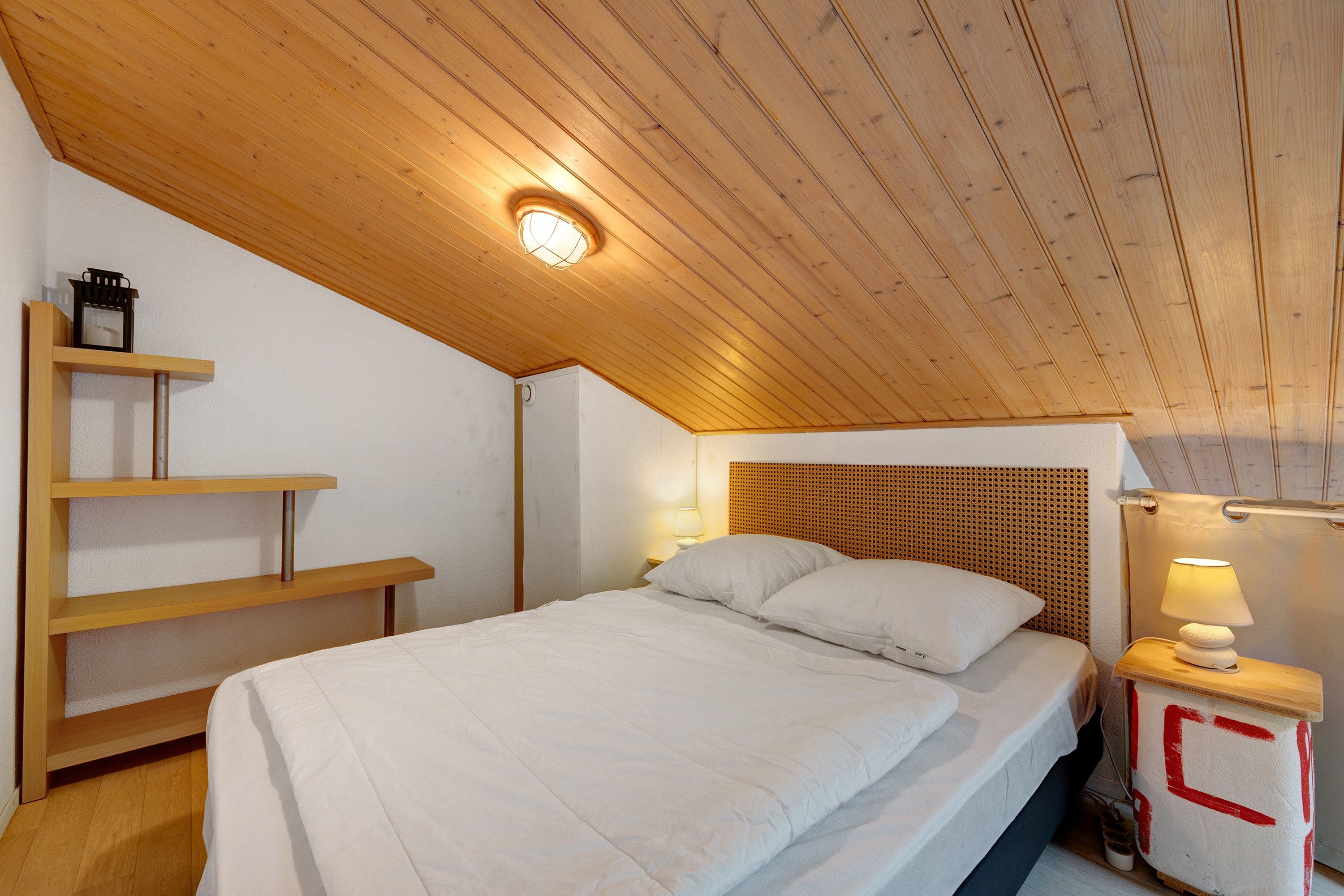 A bedroom with a wooden sloped ceiling, a bed with white bedding, a nightstand with a lamp, and a shelf with a small item on it.