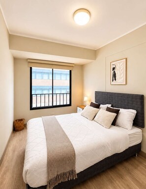 A bedroom with a large bed, a nightstand, a lamp, a framed picture on the wall, and a window with a view of buildings. - 2 dormitorios San Isidro AC y coworking (Lima)