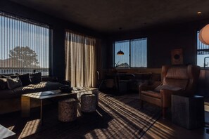 A modern living room with a sofa, armchairs, a coffee table, and a dining table. Large windows provide views of the outdoors. - AMAR By Homestead Modern (Pioneertown)
