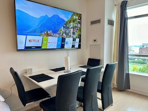 A hotel room with a large flat-screen TV mounted on the wall displaying a scenic view, a dining table with chairs, and a window with curtains. - Cozy 3 Beds Suite (Toronto)