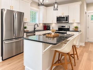 A modern kitchen with stainless steel appliances, white cabinetry, and a central island with bar stools. - The Haven at Holly Springs (Holly Springs)