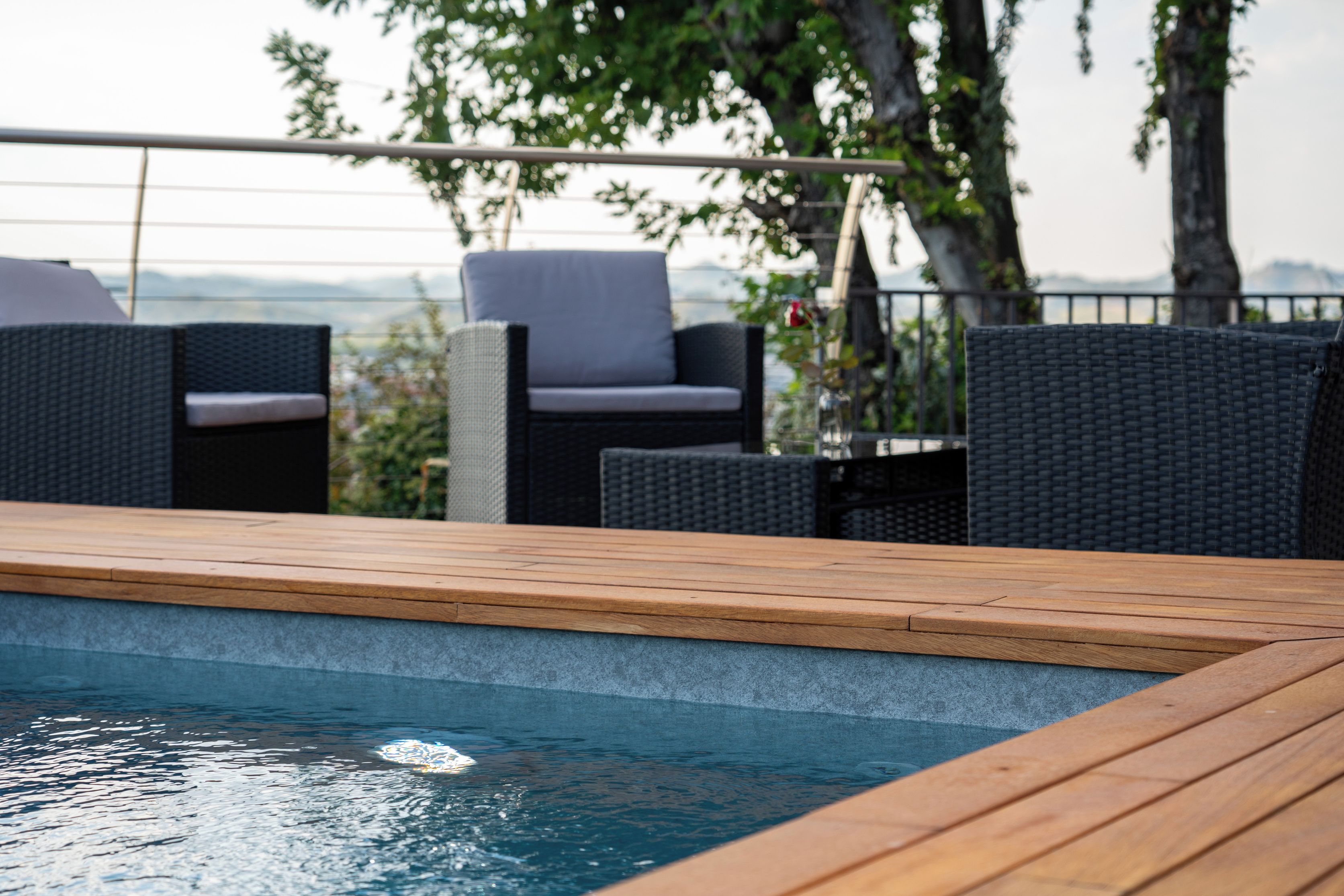 Zona junto a la piscina con terraza de madera, sillas de mimbre y vistas al paisaje.