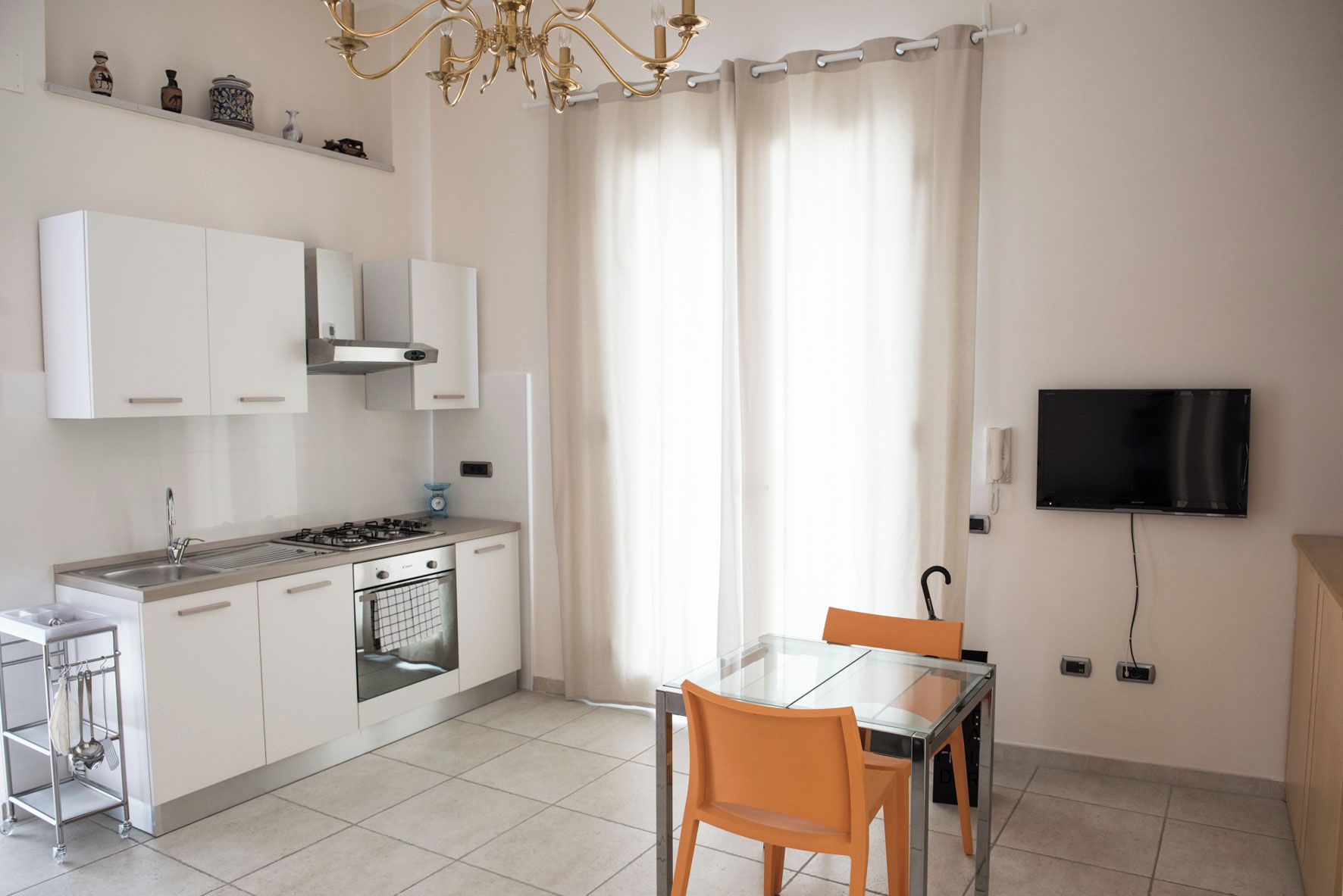 Een moderne keuken met witte kastjes, een gasfornuis en een inbouwoven. Er is een eethoek met een glazen tafel en twee stoelen. Een televisie is aan de muur bevestigd, en een kroonluchter hangt aan het plafond.