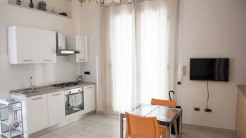 Een moderne keuken met witte kastjes, een gasfornuis en een inbouwoven. Er is een eethoek met een glazen tafel en twee stoelen. Een televisie is aan de muur bevestigd, en een kroonluchter hangt aan het plafond.