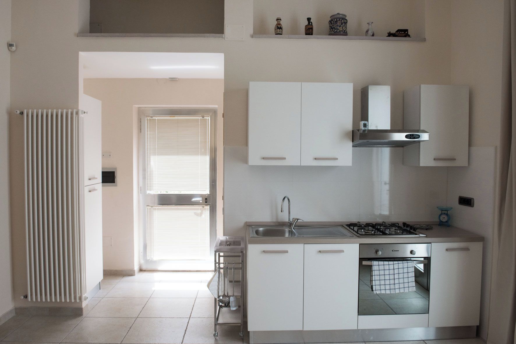 A modern kitchen with white cabinets, a stainless steel range hood, and a built-in oven. There is a window with blinds, a radiator, and a small shelf with decorative items.
