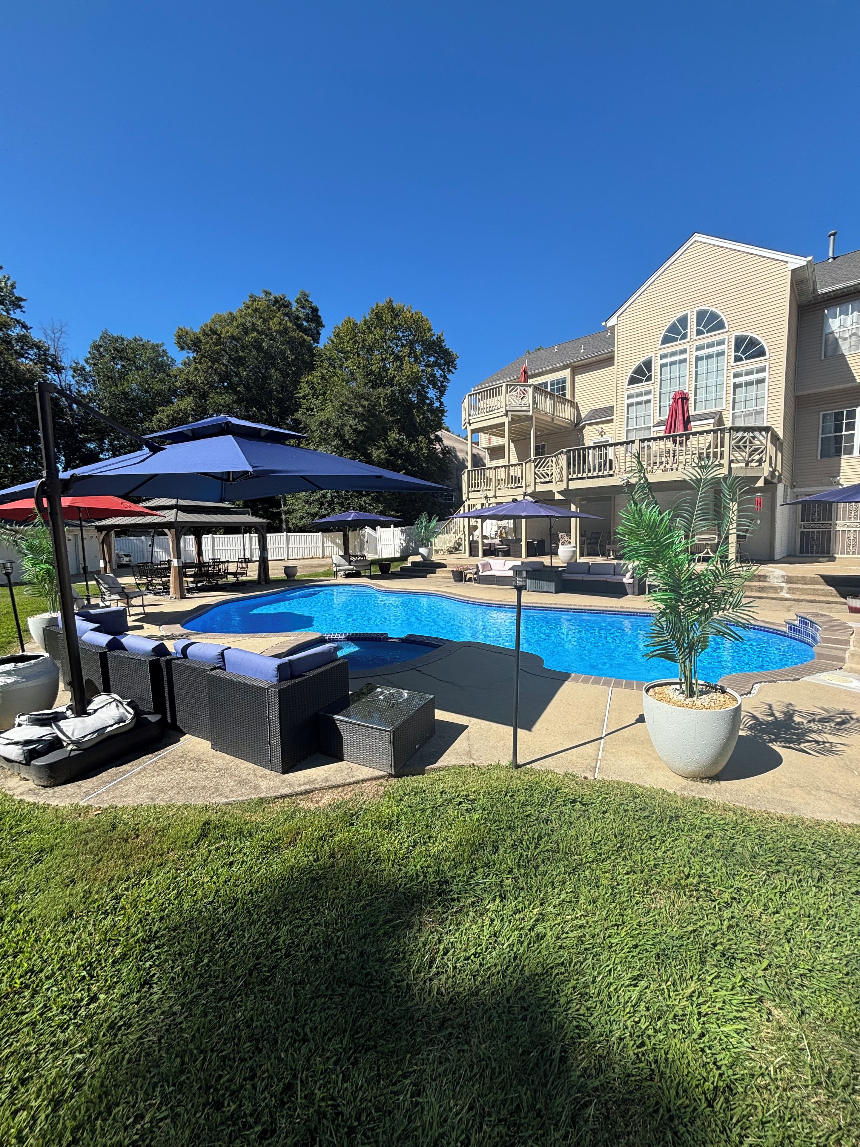 A swimming pool with lounge chairs and umbrellas, surrounded by a well-maintained lawn and a large house with a balcony.