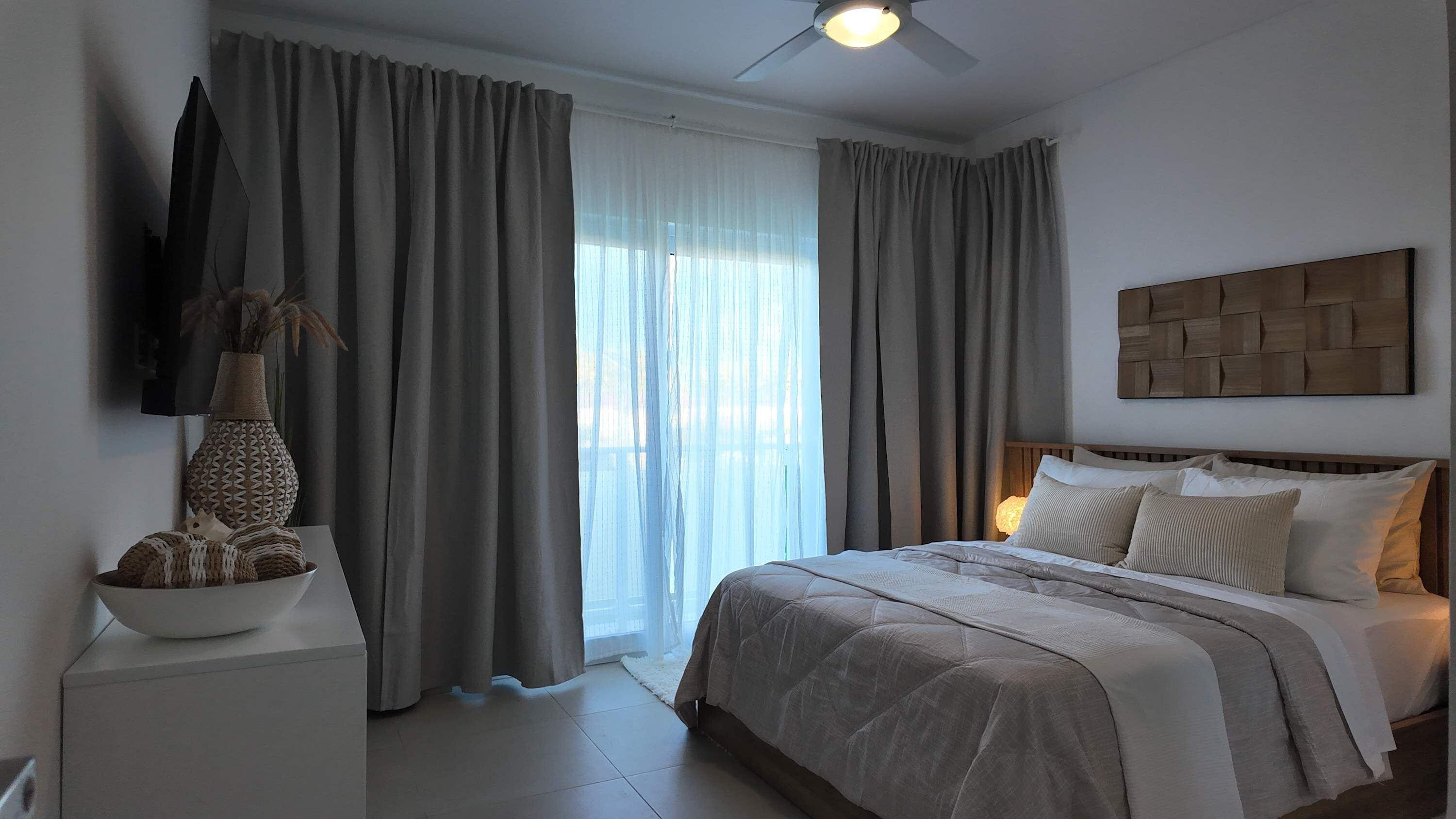 Un dormitorio con cama, cortinas, ventilador de techo, televisor montado en la pared y un jarrón decorativo.