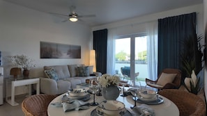 Ein helles Wohnzimmer mit einer Couch, einem Esstisch für zwei Personen und einem großen Fenster mit Vorhängen.