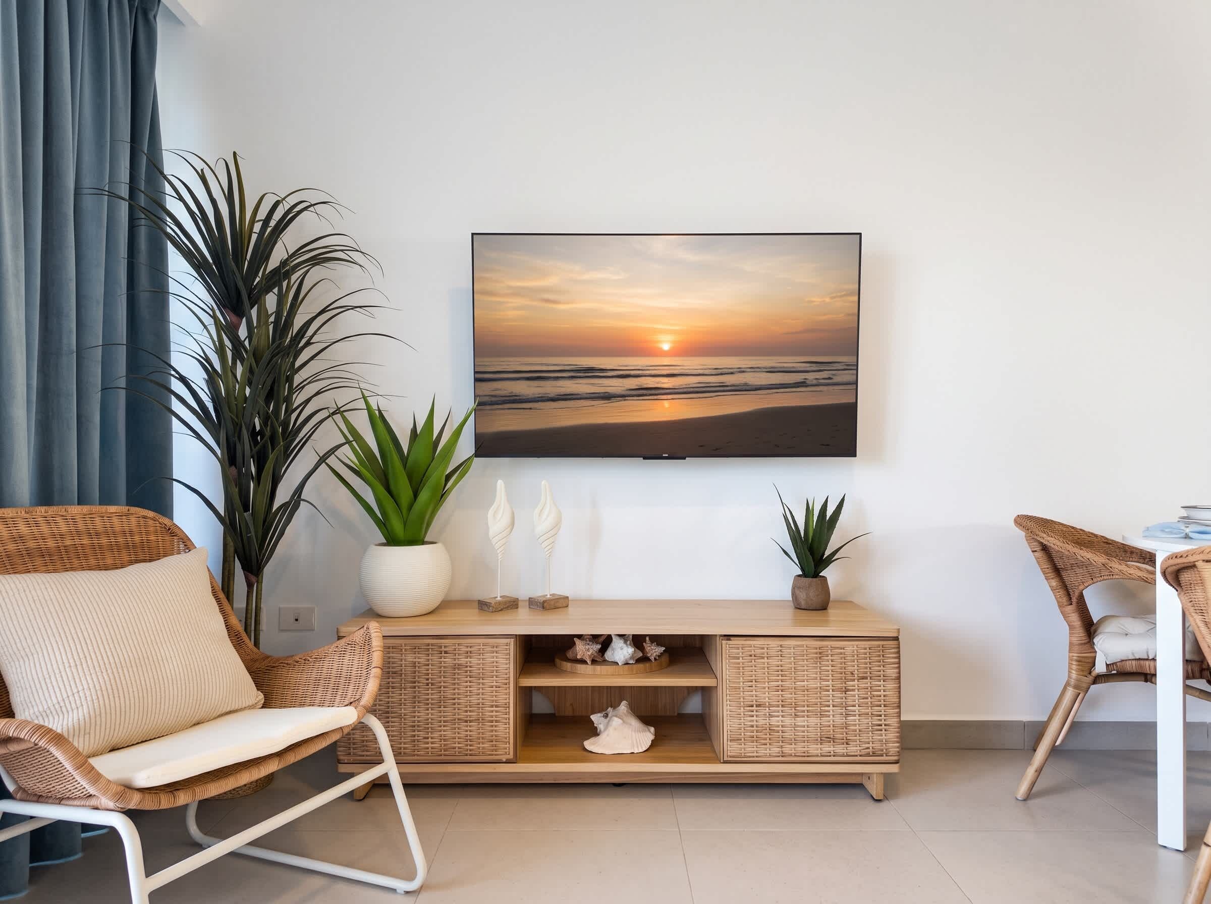 A living room with a flat-screen TV mounted on the wall, a wicker sofa, a small wooden side table with decorative items, and a potted plant.